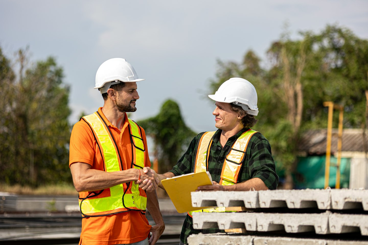 Two construction workers in hard hats shake hands near concrete slabs.